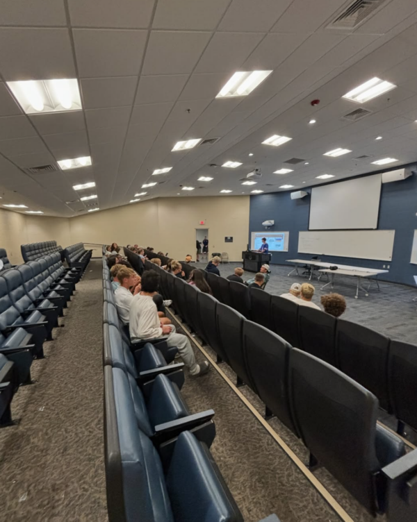 People sit in rows of seats in a lecture hall, facing a presenter at the front who is speaking near a projector screen and whiteboards during an FCA (Fellowship of Christian Athletes) event.